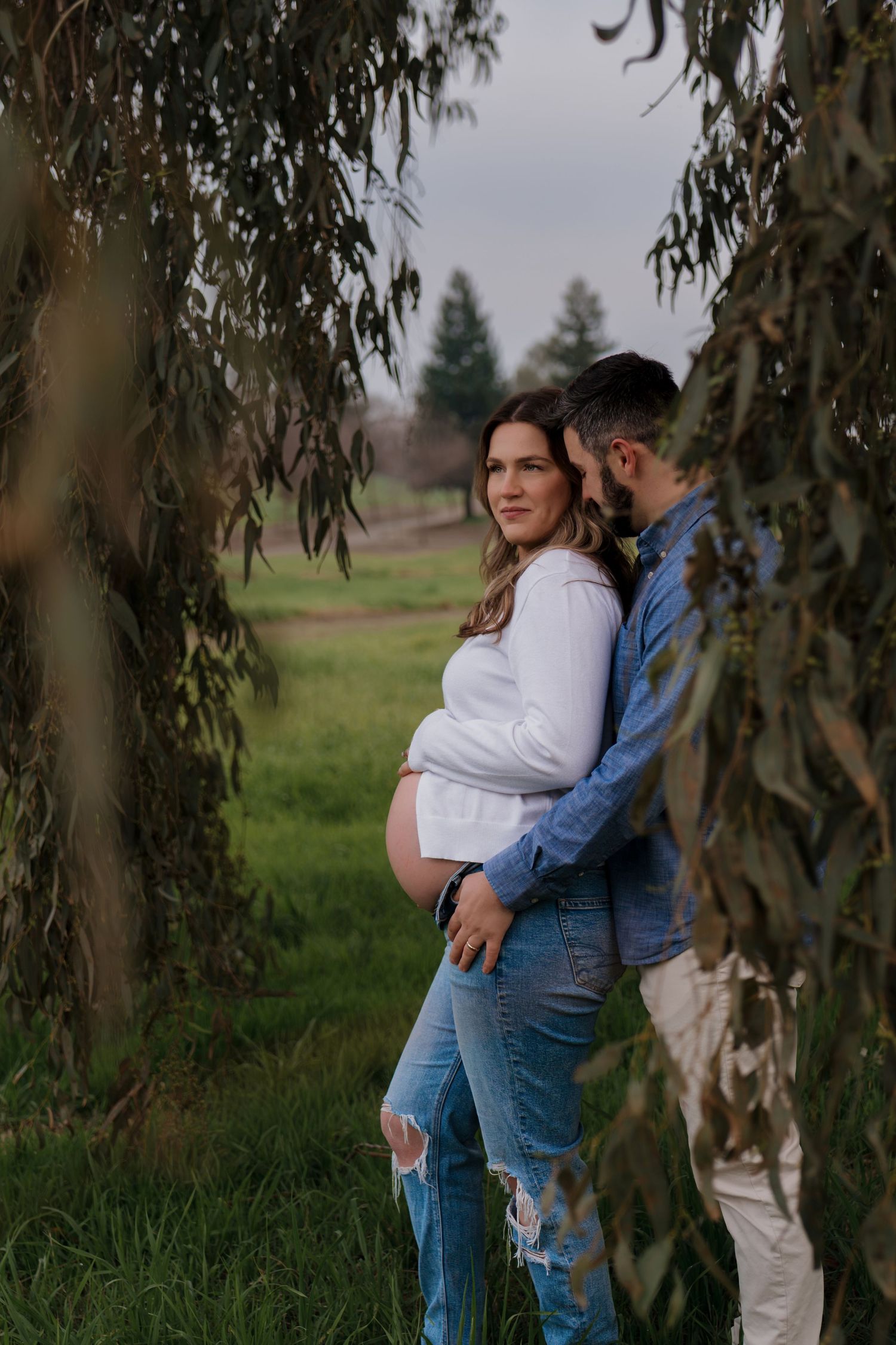 Intimate maternity photos captured under willow trees with a couple in casual denim and white attire.