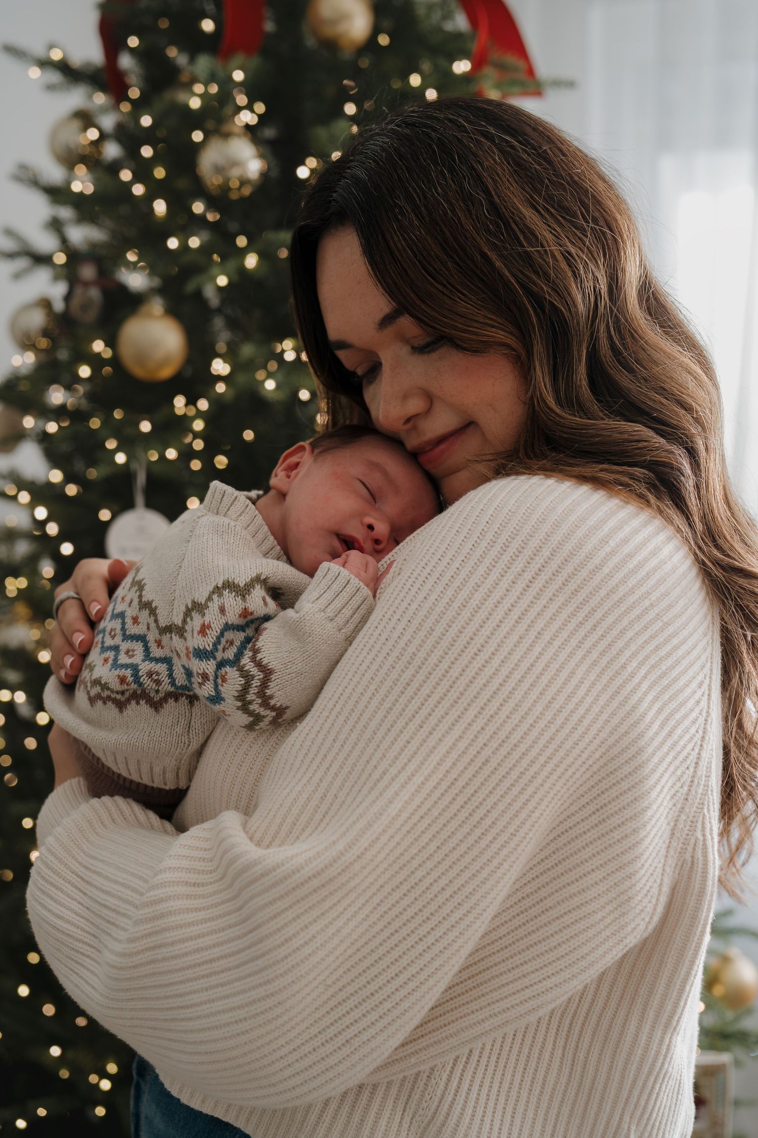 A tender moment between parent and newborn baby in front of a decorated Christmas tree with twinkling lights and ornaments.