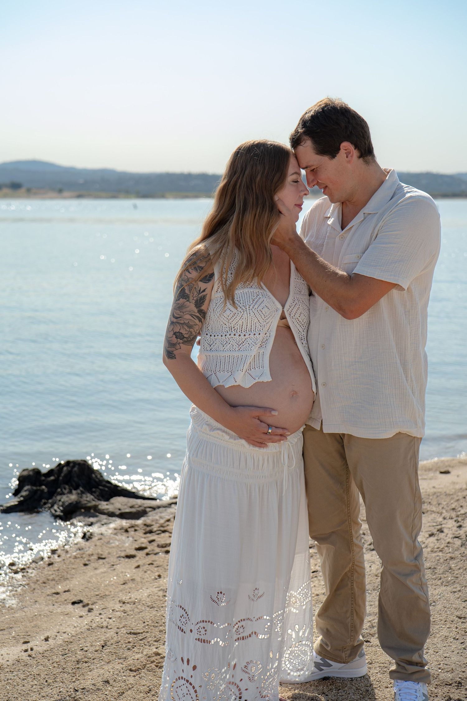 Couple in white outfits sharing romantic moment on rocky beach.