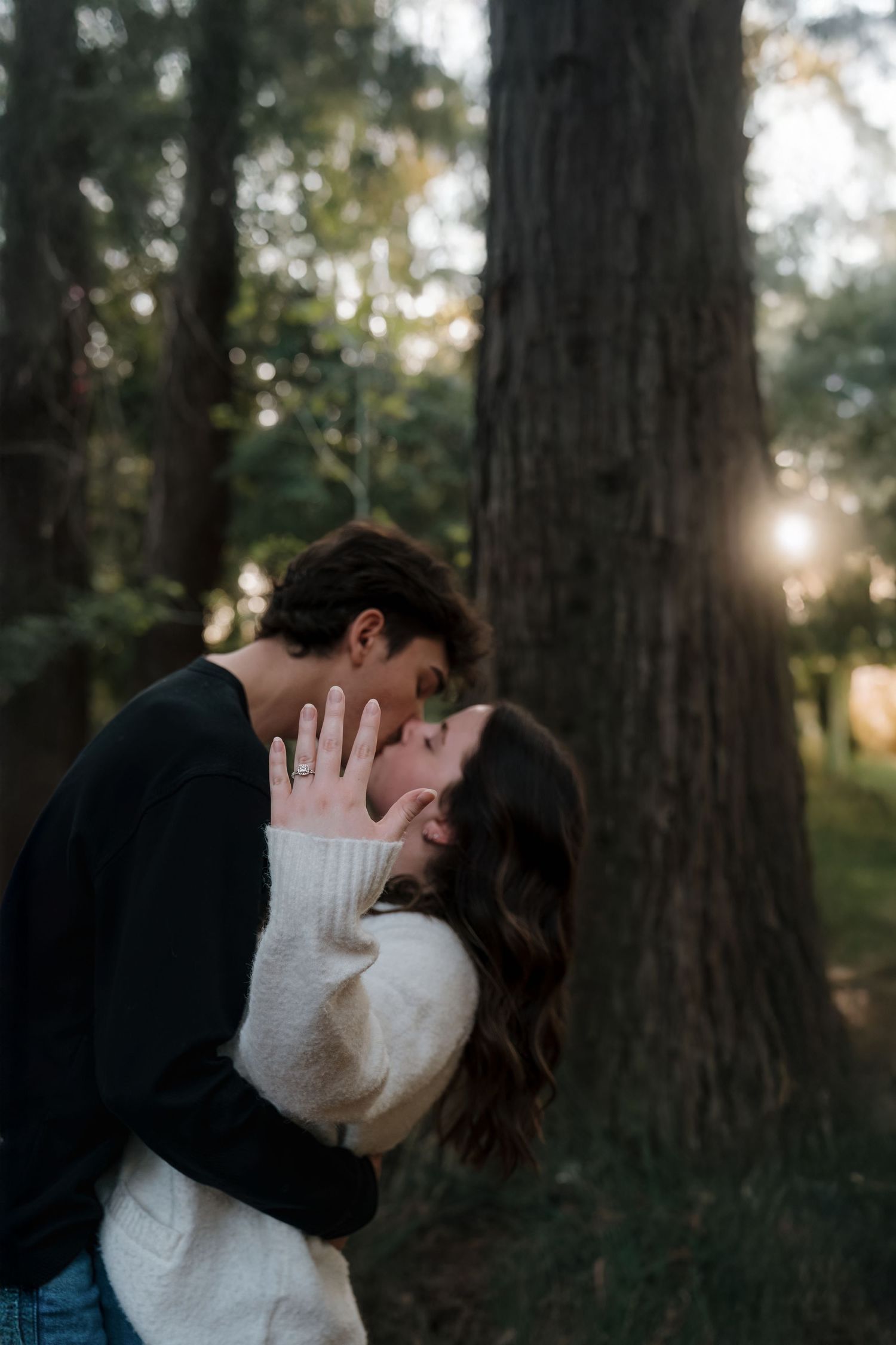 Couple sharing a romantic kiss in a sunlit forest, woman in white sweater showing engagement ring.
