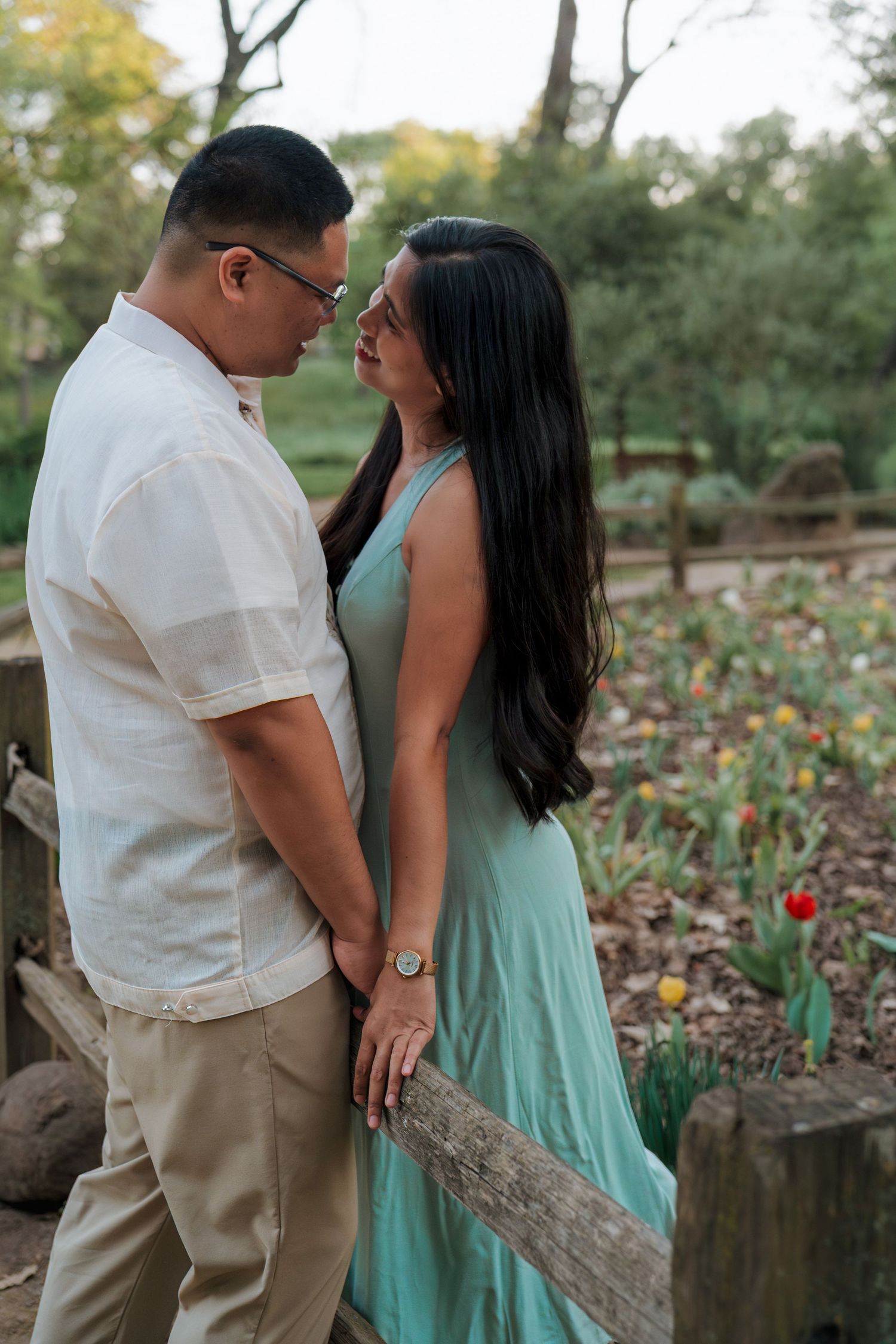 Couple in a garden holding hands and gazing at each other, woman in teal dress, man in white shirt, tulips in background.