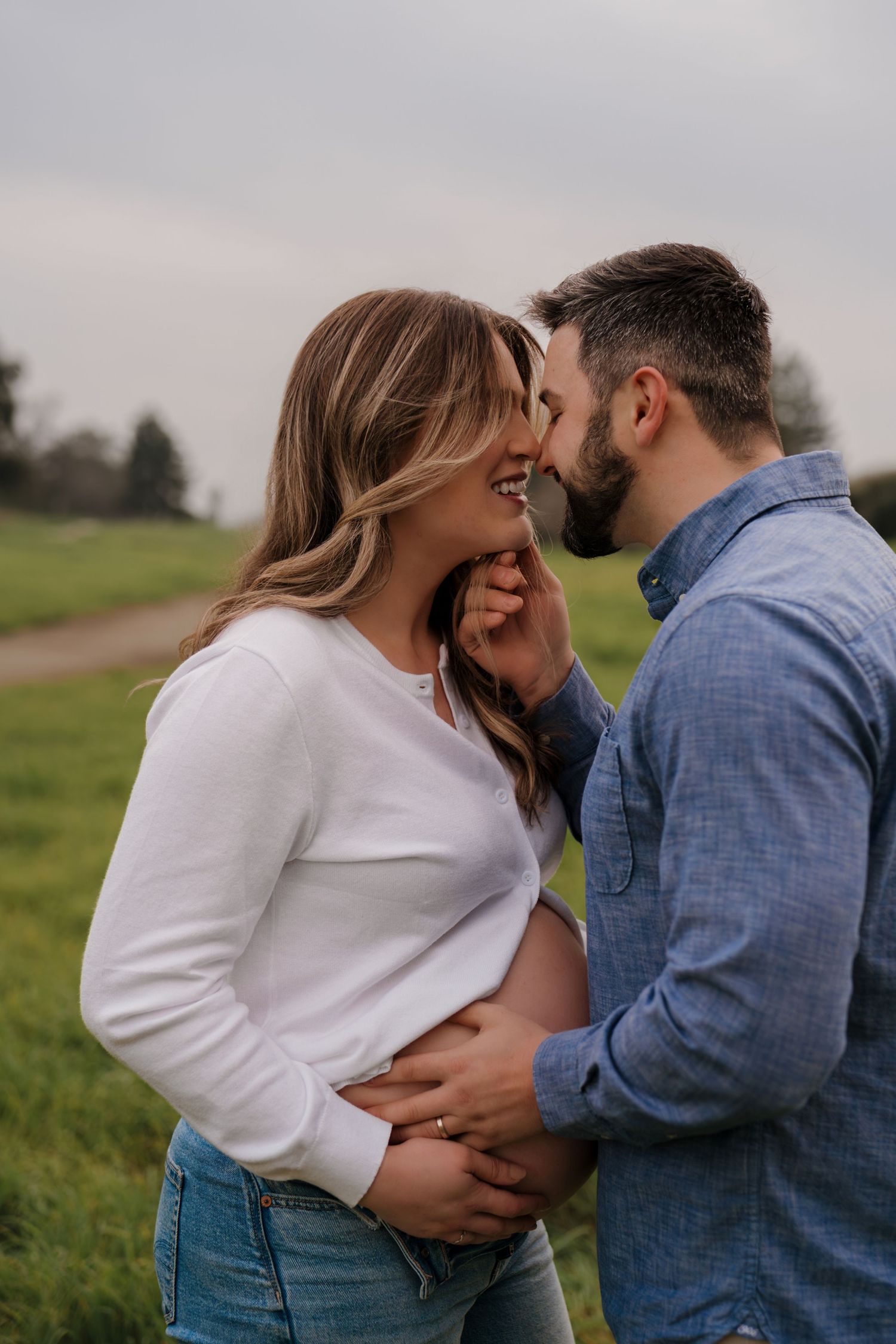 A romantic couple in denim and white embrace in a grassy field during an outdoor photo session.