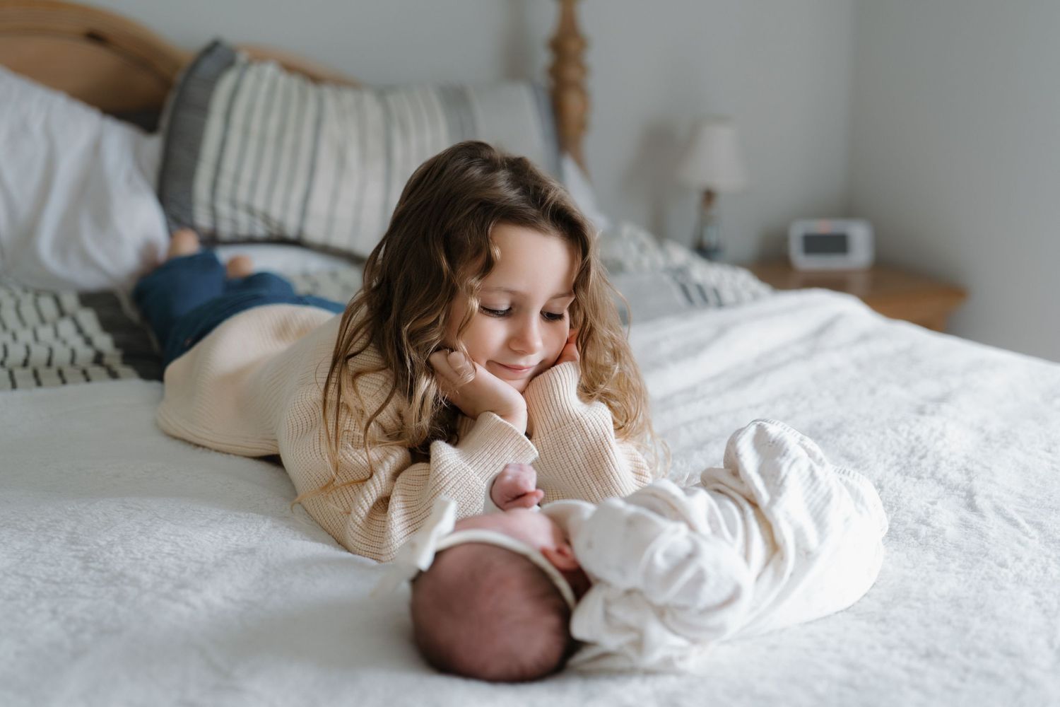 A child in white clothing plays and rolls around on a cozy white bed with natural lighting.