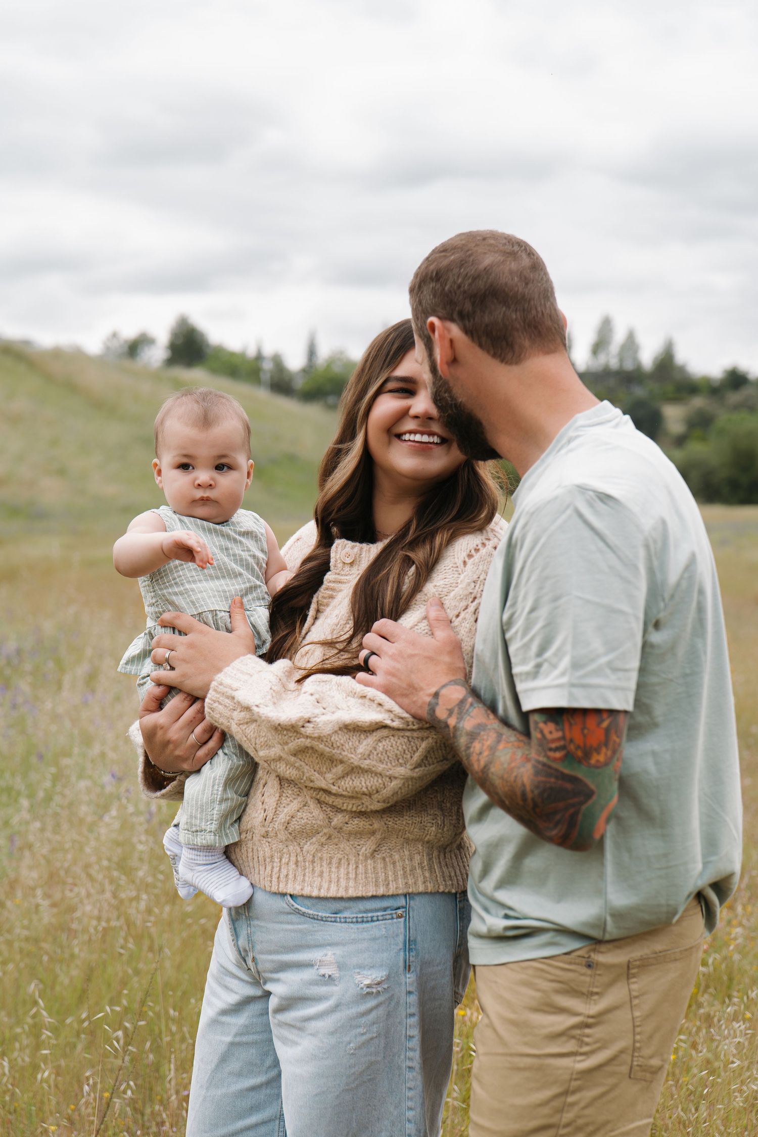 Happy young couple with tattoos holding their baby in a scenic green meadow under cloudy skies.