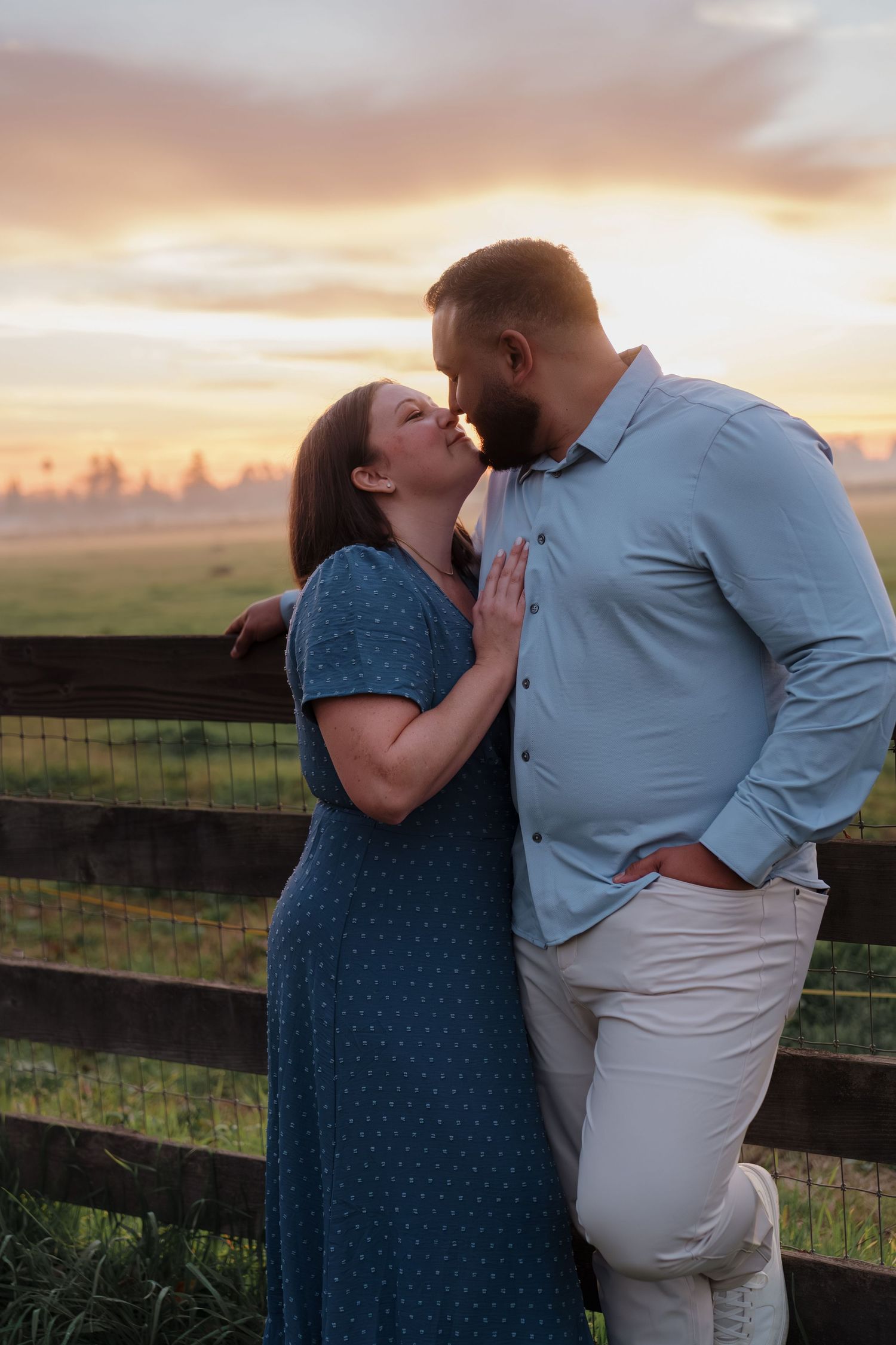 A couple shares romantic moments at sunset by a wooden fence in a rural setting with golden skies in the background.