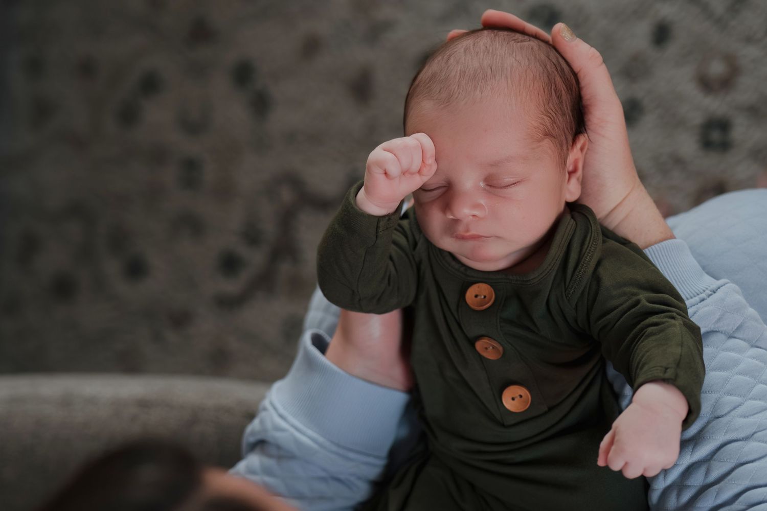 A sleeping newborn baby wearing a dark outfit lies on a light blue blanket while being gently cradled.