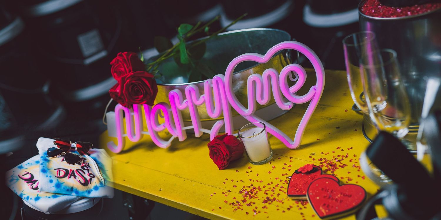 Pink neon 'Marry Me' sign displayed on yellow surface with red roses, confetti, and champagne glasses for proposal setup.