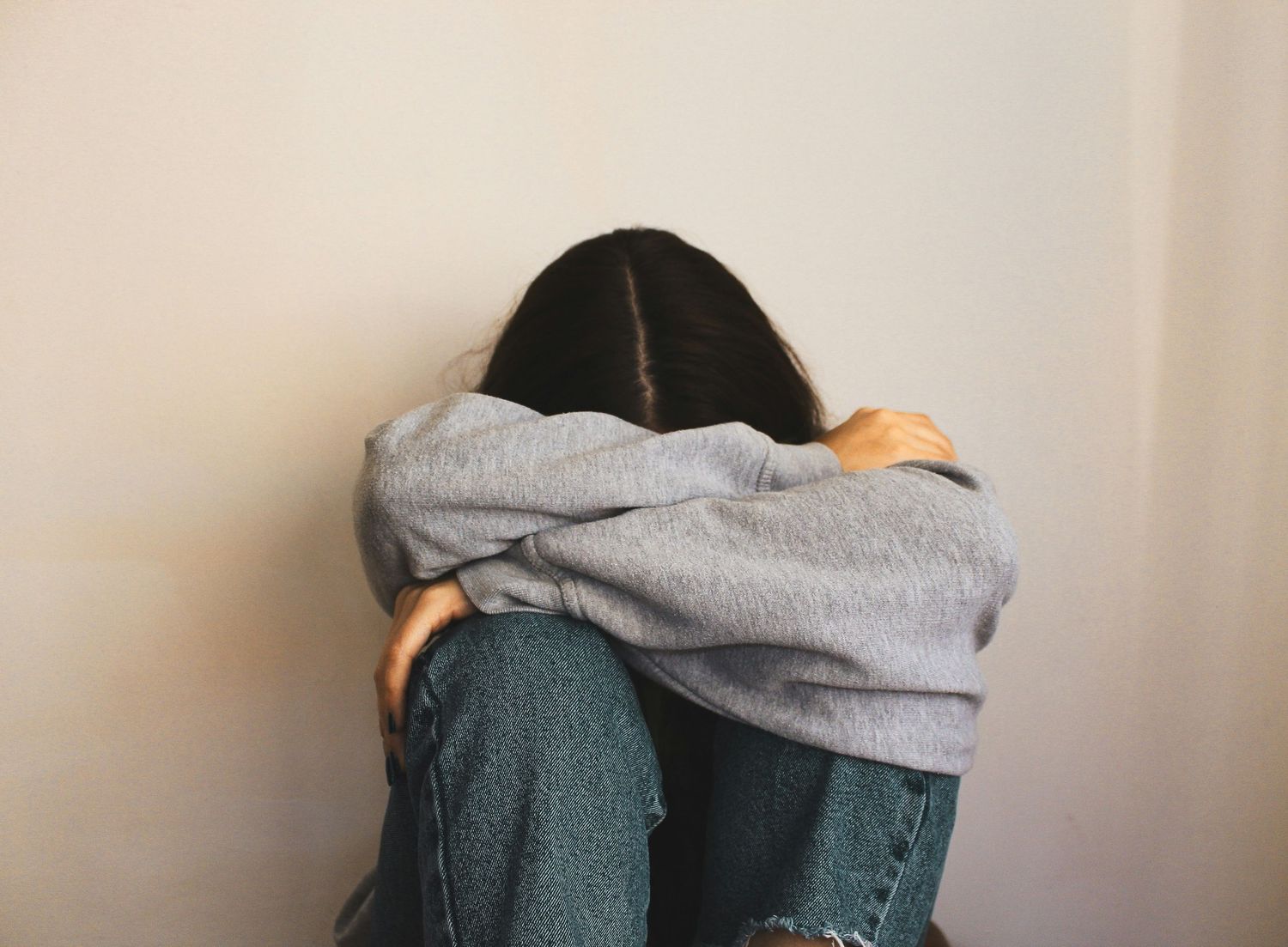 A person in jeans and a gray hoodie sits curled up with their knees to their chest in a corner upset about her wedding photos.