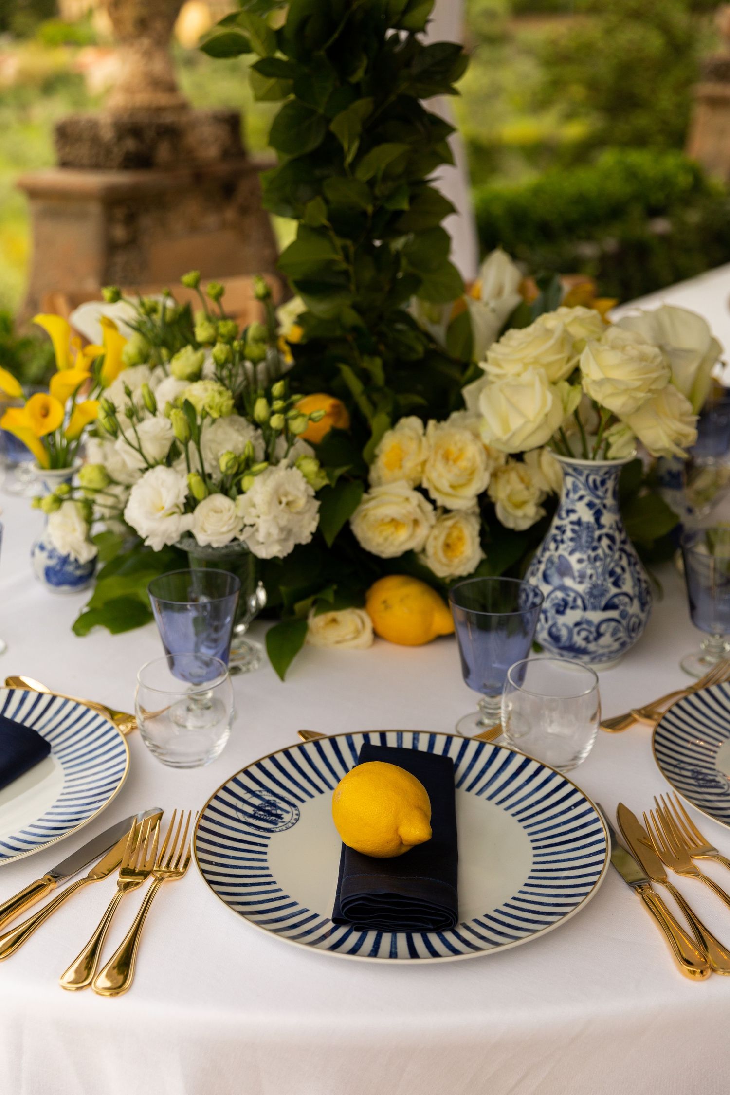 Elegant table setting with blue and white striped plates, gold cutlery, and white floral centerpieces with yellow accents.