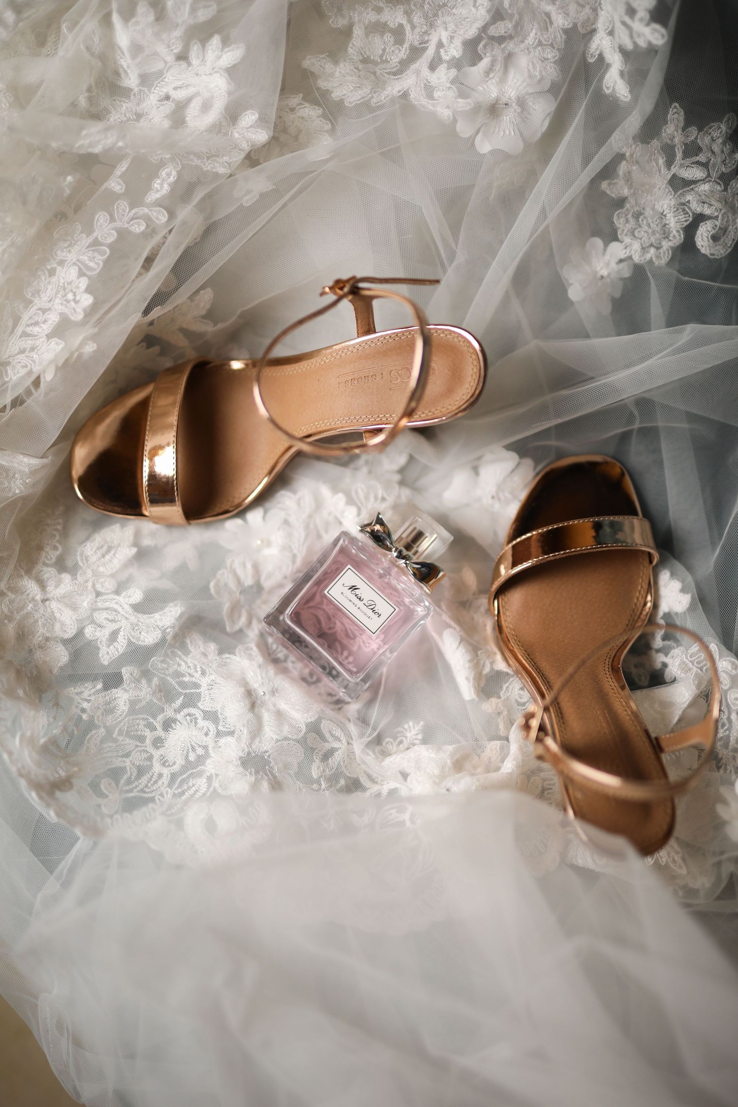 Elegant rose gold bridal sandals and perfume bottle arranged on delicate white lace veil.