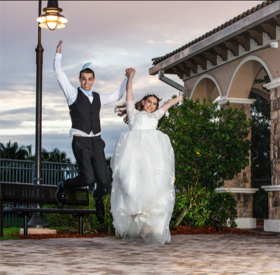 A romantic wedding couple jumps up together in an elegant outdoor terrace with arched architecture in the background.