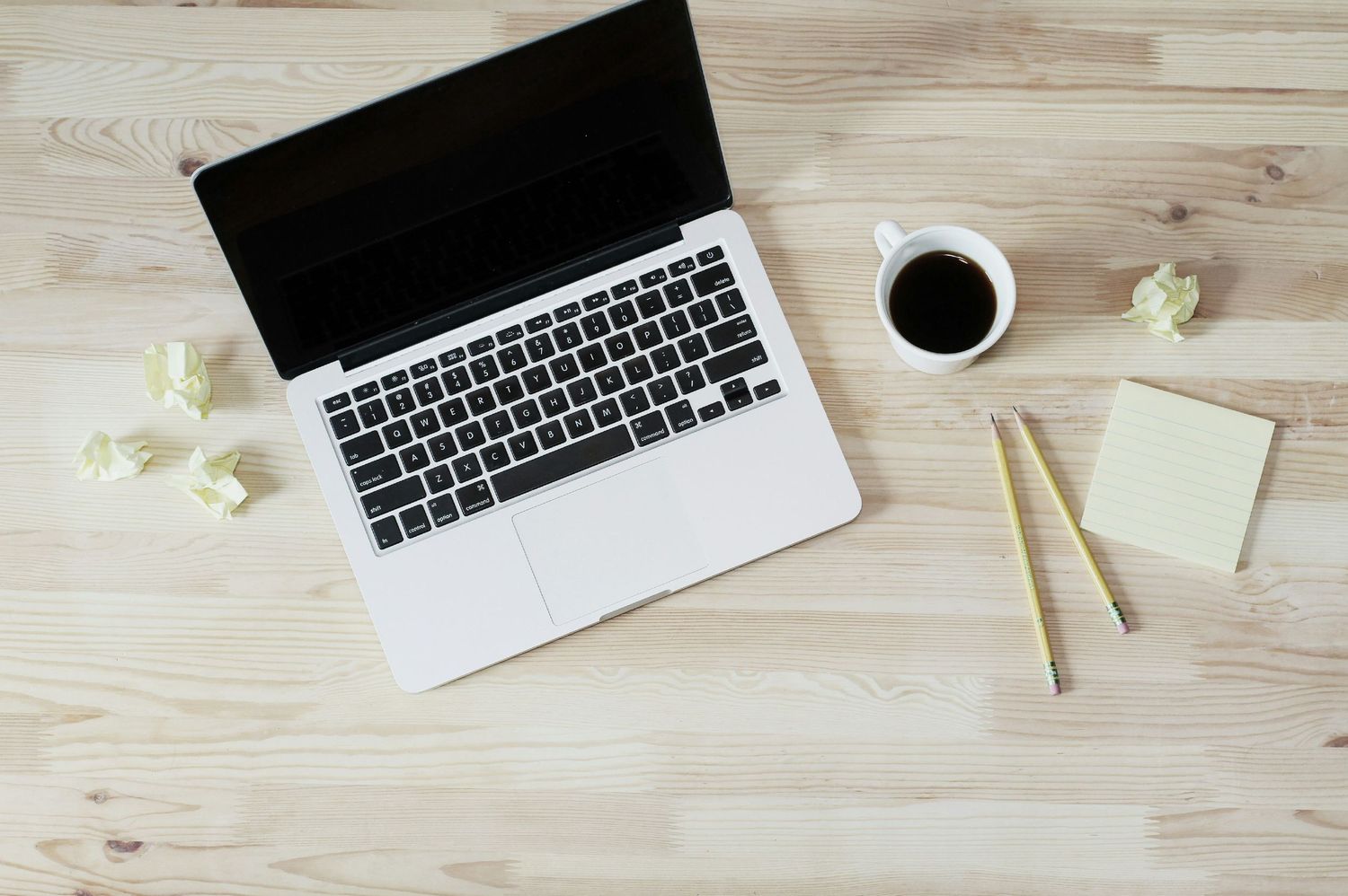 Laptop and coffee cup on light wooden desk with crumpled paper and scattered office supplies.