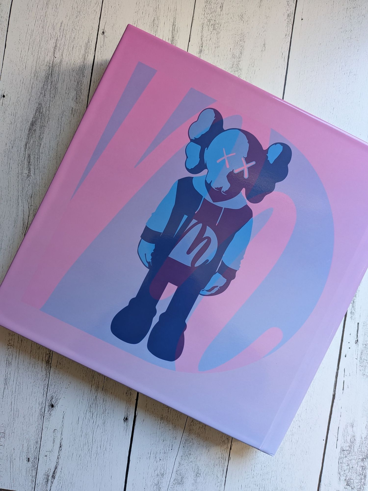 Pink KAWS hardcover art book featuring a blue companion figure graphic on a white wooden surface.