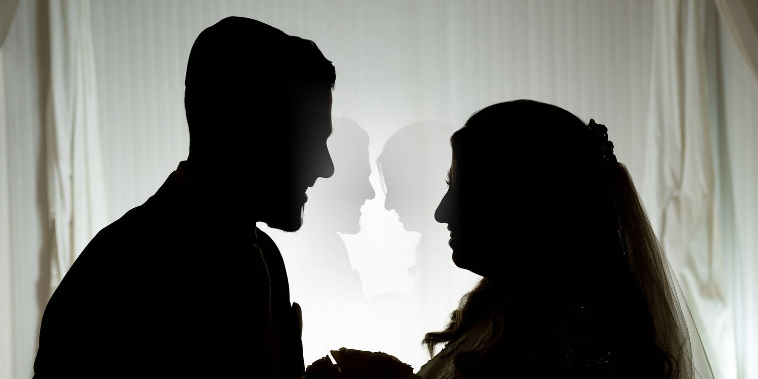 Silhouettes of a bride and groom standing against a bright white background in a dramatic composition.