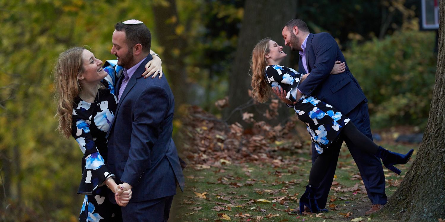 A couple in formal engagement attire embraces while standing outdoors in an autumn setting with falling leaves.