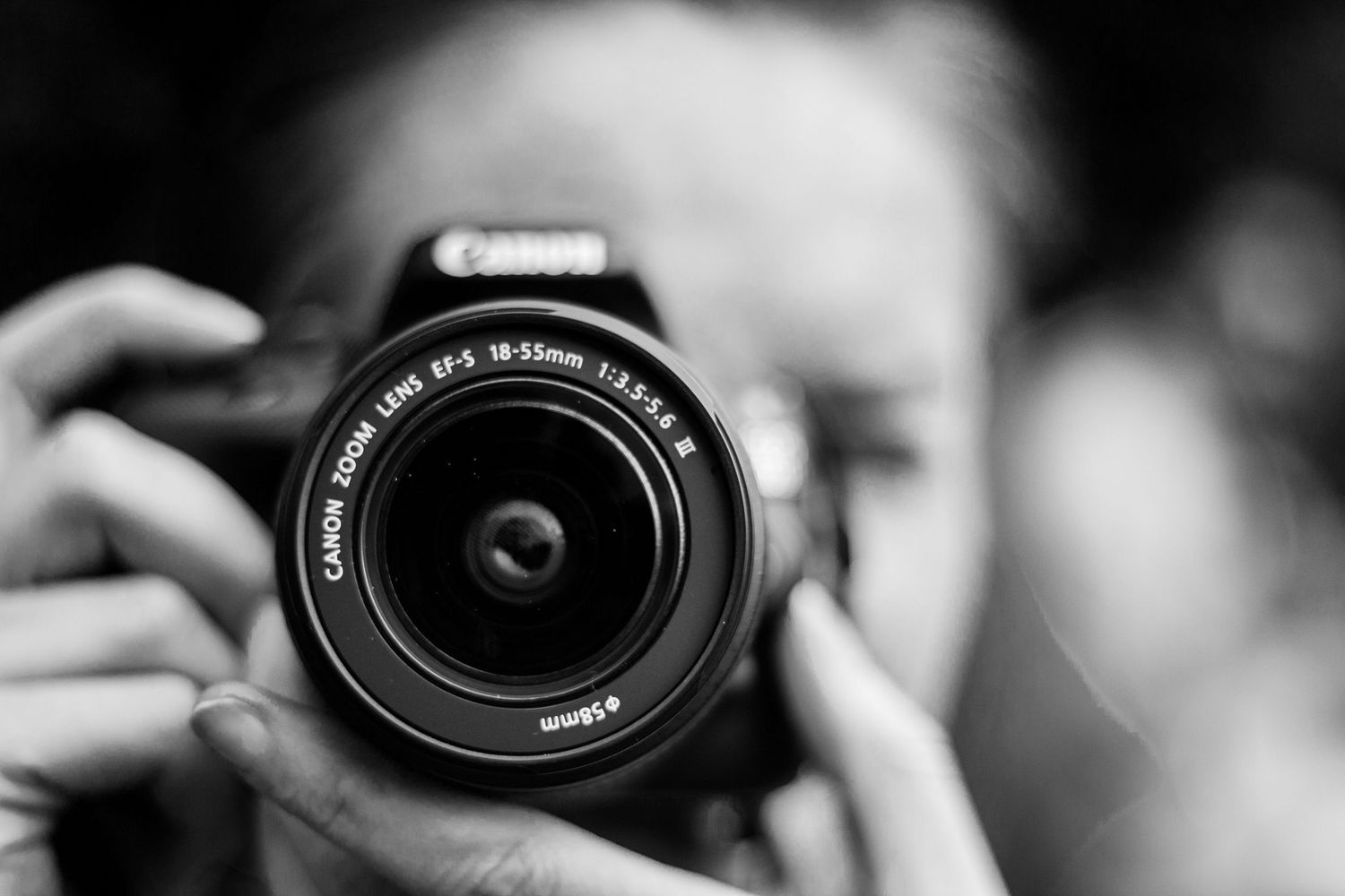 A close-up black and white photo of a Canon camera lens being held up to take a photograph.