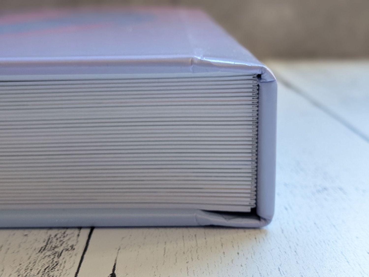 Close-up of a thick hardcover book spine with ribbed binding in soft lavender gray, resting on a white wooden surface.