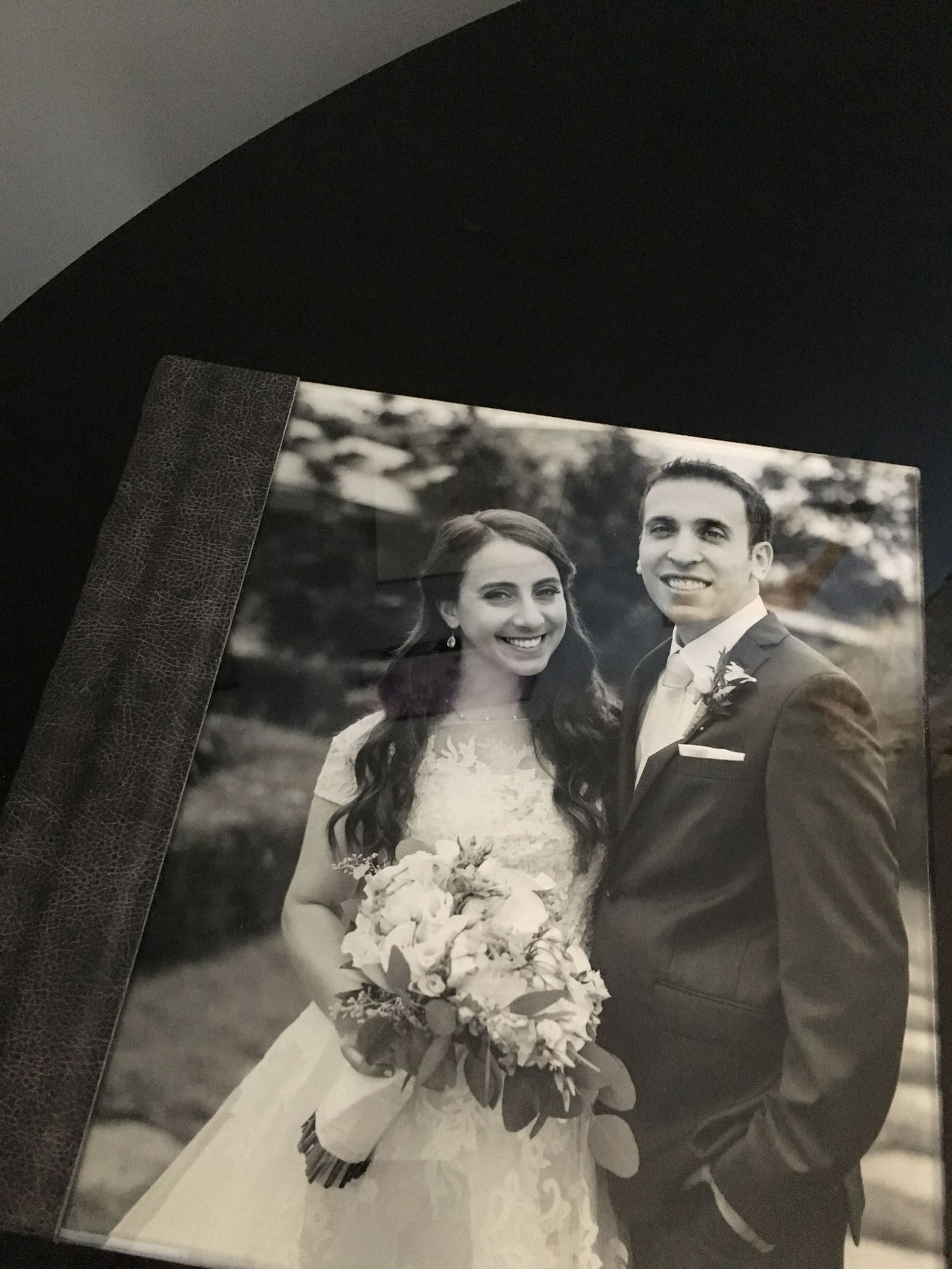 A black and white acrylic wedding photo album showing a bride in a lace gown holding a floral bouquet next to a groom in a dark suit.