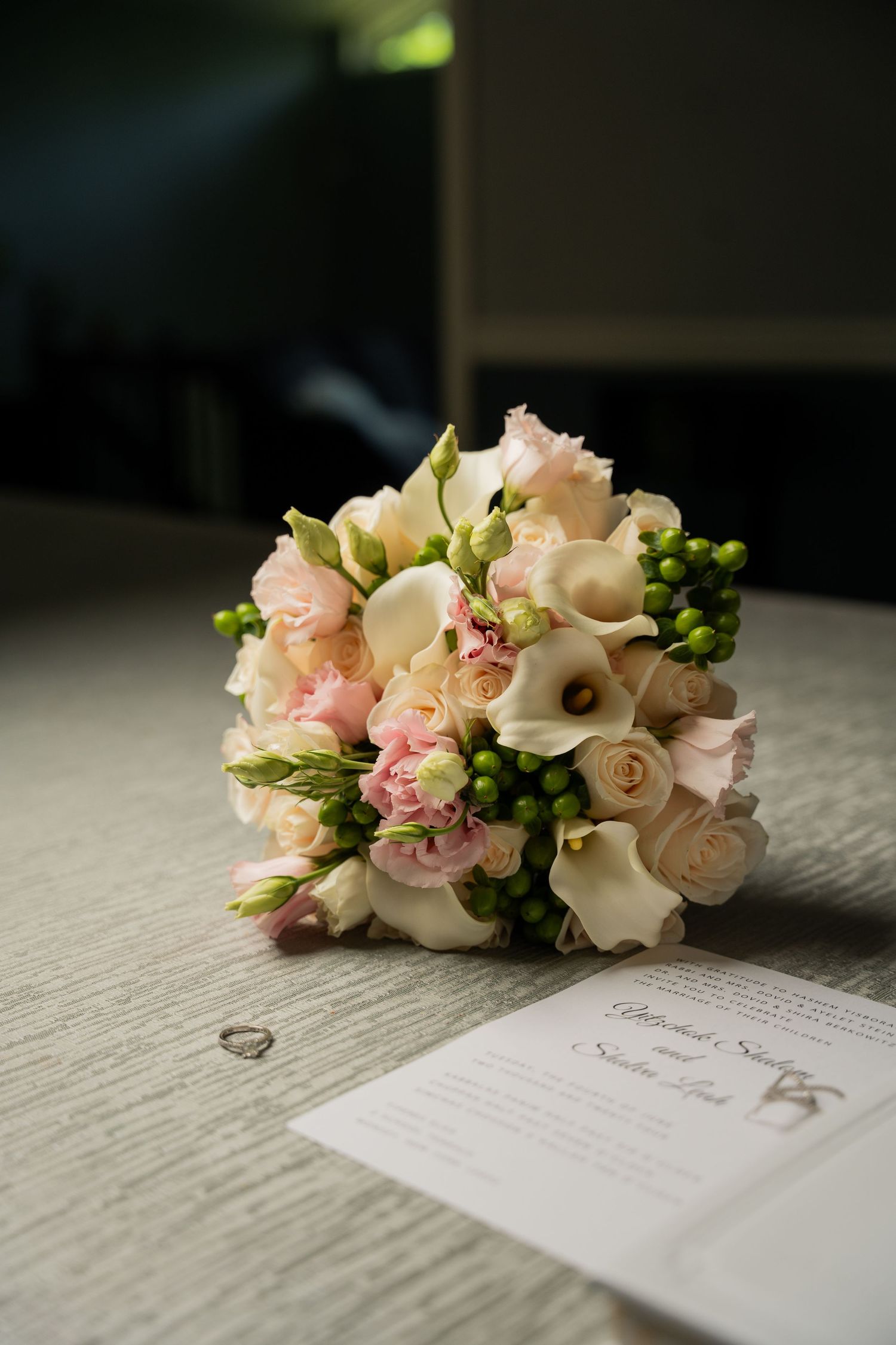 Elegant pink roses and white calla lily bouquet rests beside wedding invitation cards on textured fabric surface.