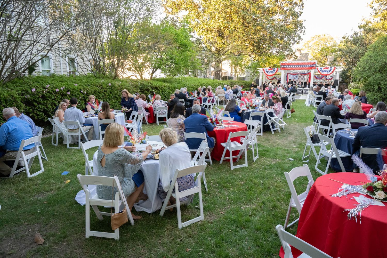 Outdoor garden party with guests seated at white chairs and red-clothed tables on a lawn near a red pavilion.