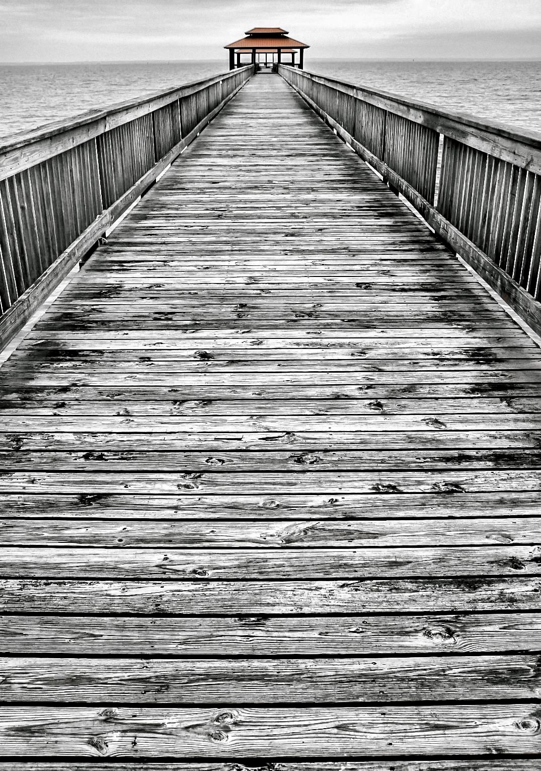 Black and white photograph of a wooden pier extending over calm waters with a gazebo at the end.