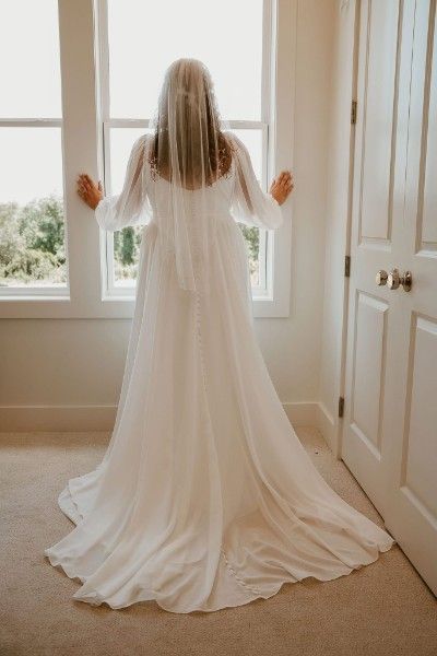 Bride in flowing white wedding gown and veil stands at window in sunlit room, seen from behind.
