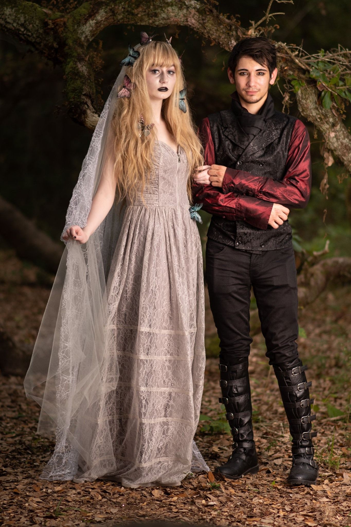 A gothic fantasy styled couple poses in a wooded setting wearing a flowy vintage dress and dark period attire.