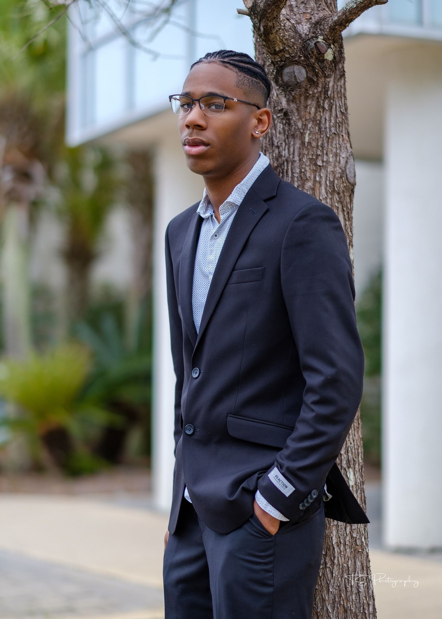 Professional headshot of young man in navy suit leaning against tree in outdoor setting.
