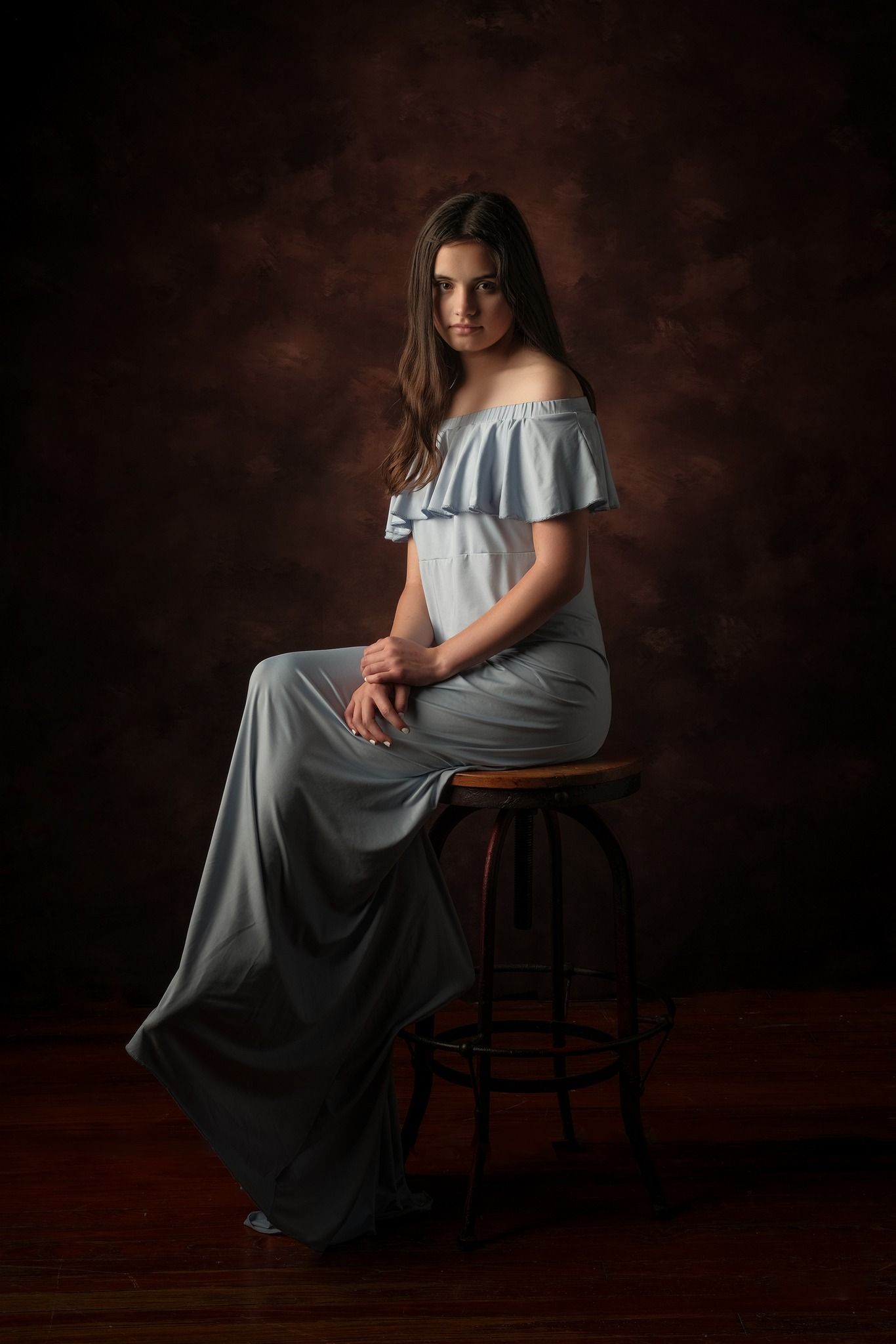 Romantic portrait in flowing off-shoulder blue dress seated on dark wooden stool.
