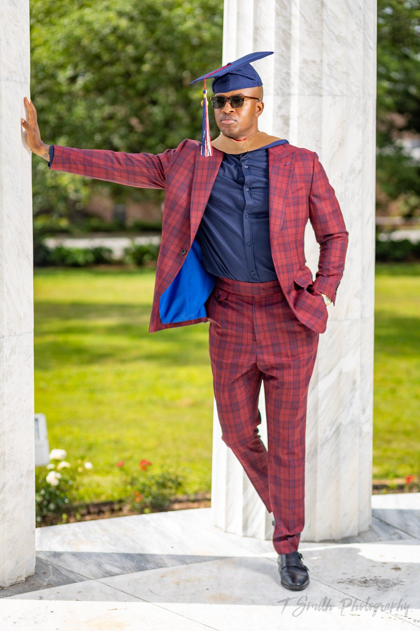 Graduate in plaid burgundy suit poses confidently against white pillars outdoors.