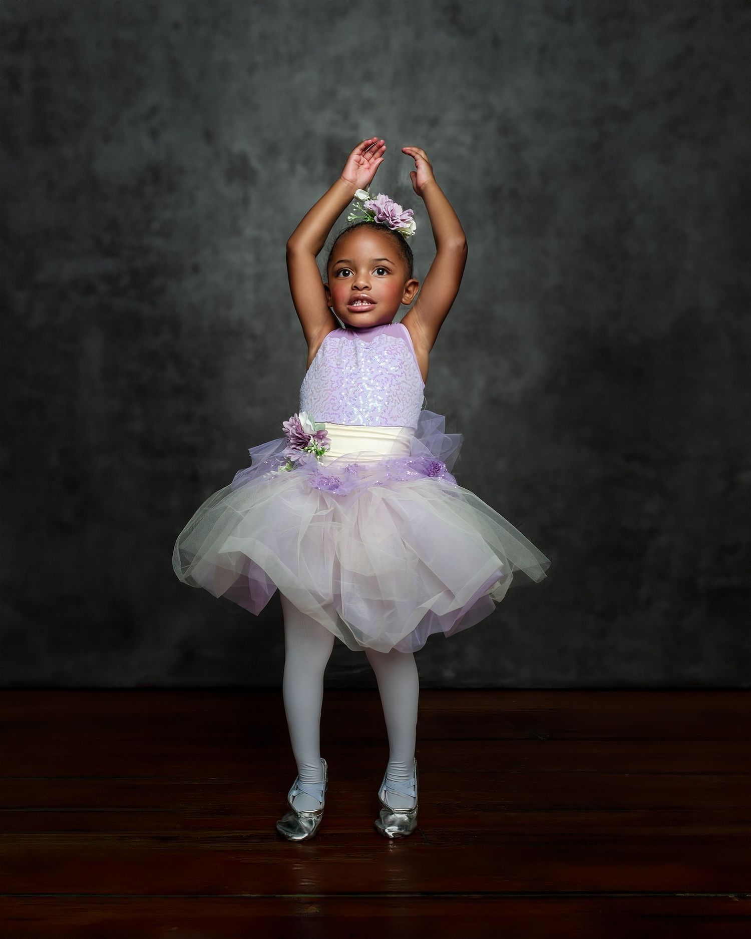 Young dancer in lavender tutu dress and crown striking ballet pose against dark backdrop.