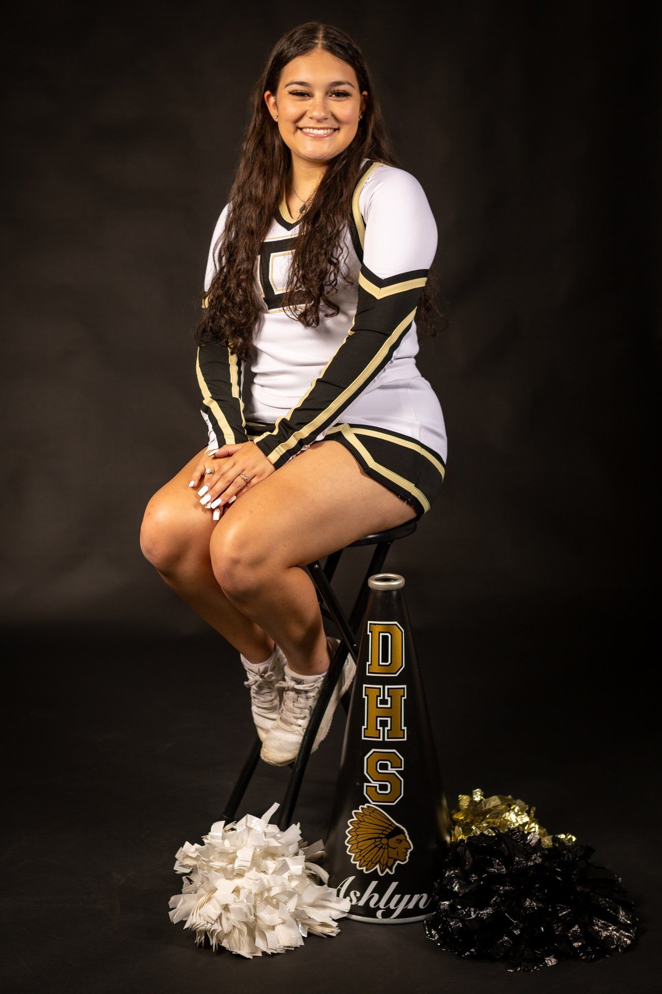 Cheerleader portrait in white and gold uniform with pom poms against dark background.