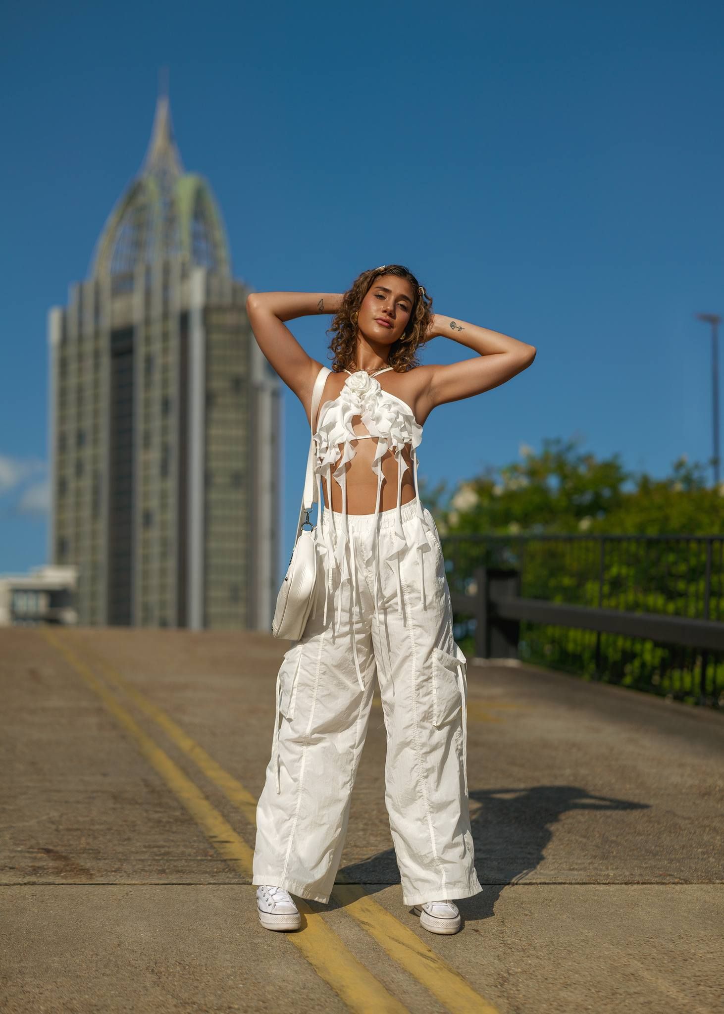 Fashion model poses in white cargo pants and crop top against urban architecture with tall building.