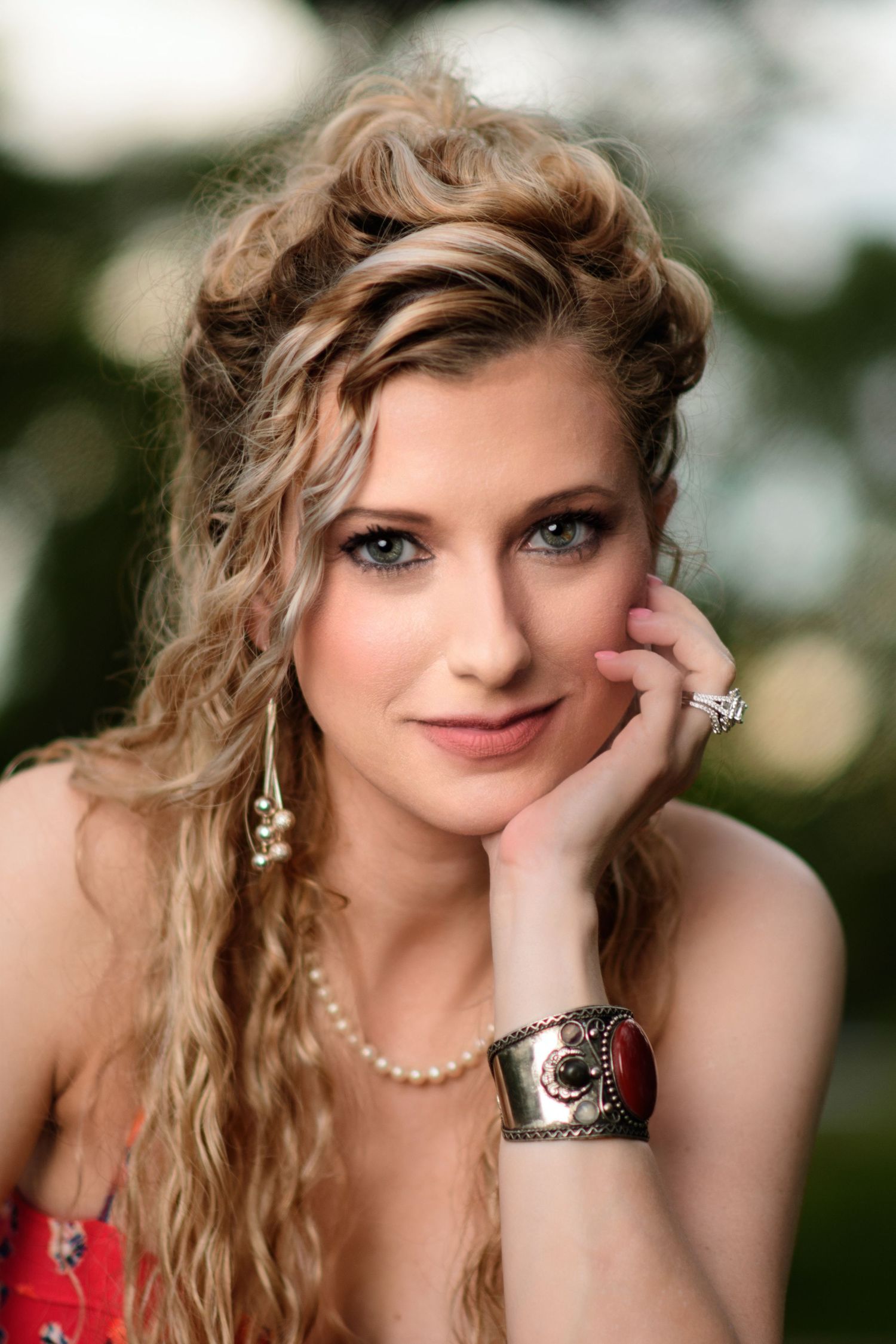 Portrait with wavy blonde hair styled up, wearing pearl necklace, dangling earrings, and decorative cuff bracelet in outdoor setting.