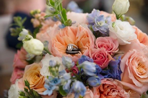 Wedding rings nestled among pastel roses and delphinium in a romantic bridal bouquet.