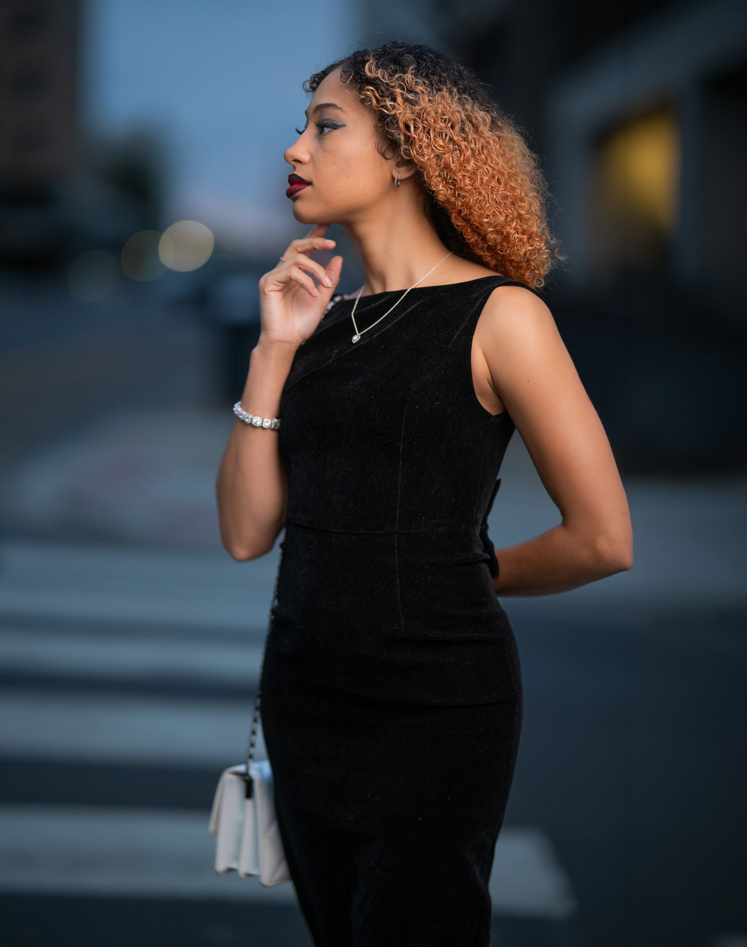 A stylish young woman in a black sleeveless dress poses outdoors at dusk, holding a white purse with curly auburn hair.