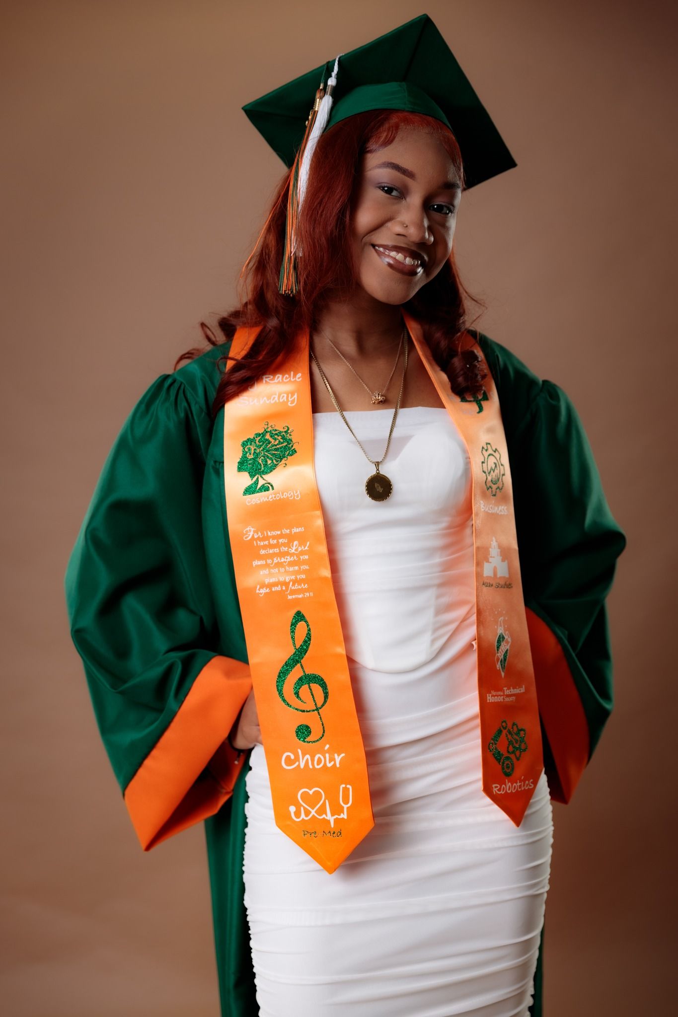 Graduate in green cap and gown with orange sash celebrating academic achievement.