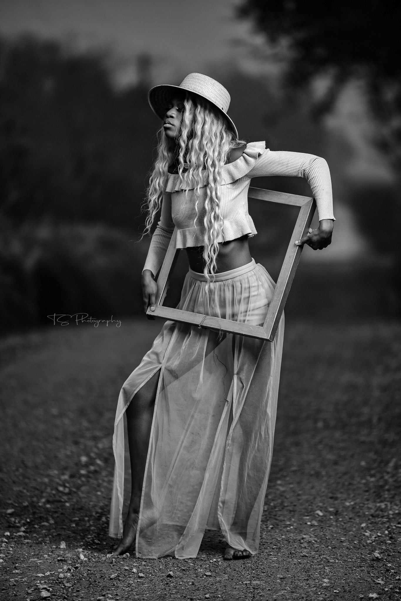 Artistic black and white portrait with model in flowing dress holding picture frame outdoors.