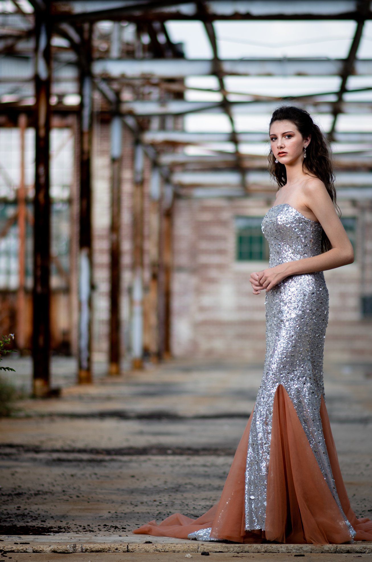 Glamorous silver sequin evening gown with thigh slit photographed in industrial setting with dramatic lighting.