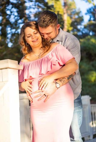 Maternity photo of couple embracing in pink dress and grey shirt outdoors.