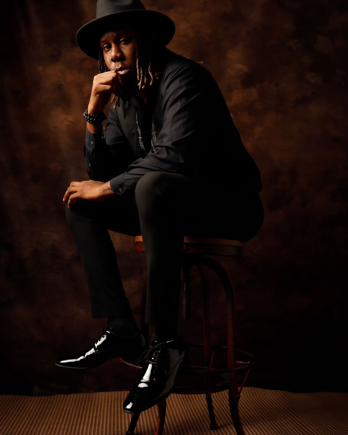 Dramatic low key portrait in black outfit and hat sitting on wooden stool.
