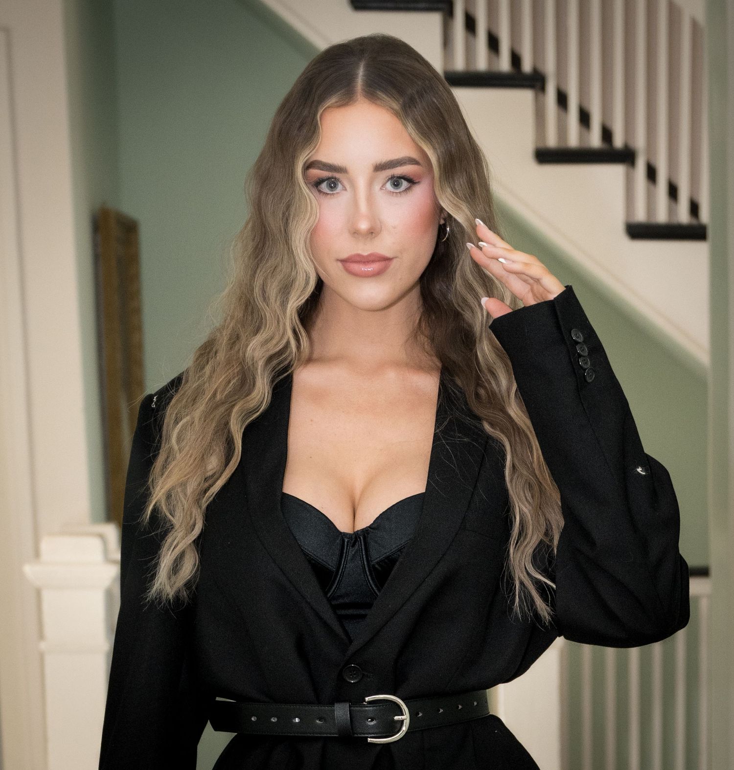 A woman in a black blazer with long wavy blonde hair poses against a staircase background.