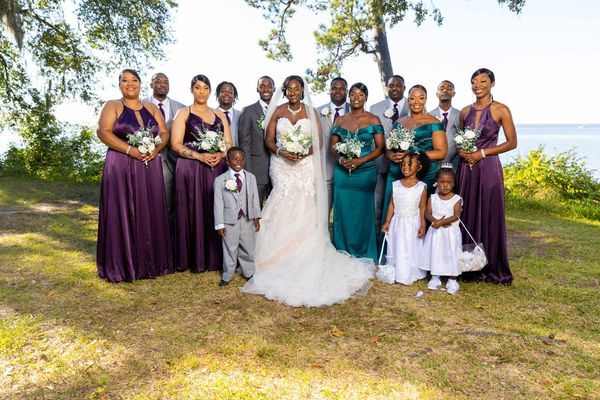 Wedding party in purple and teal dresses poses outdoors by waterfront.