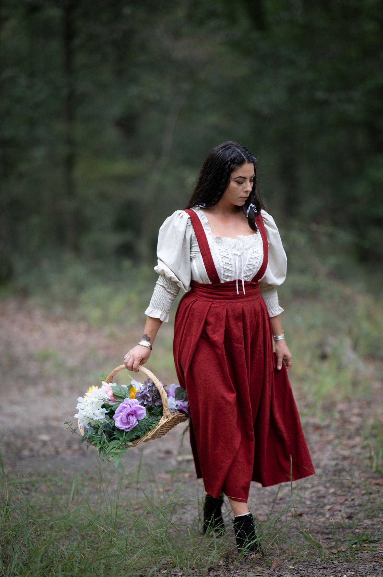 Person wearing red pinafore dress and white blouse holding flower basket in forest setting.