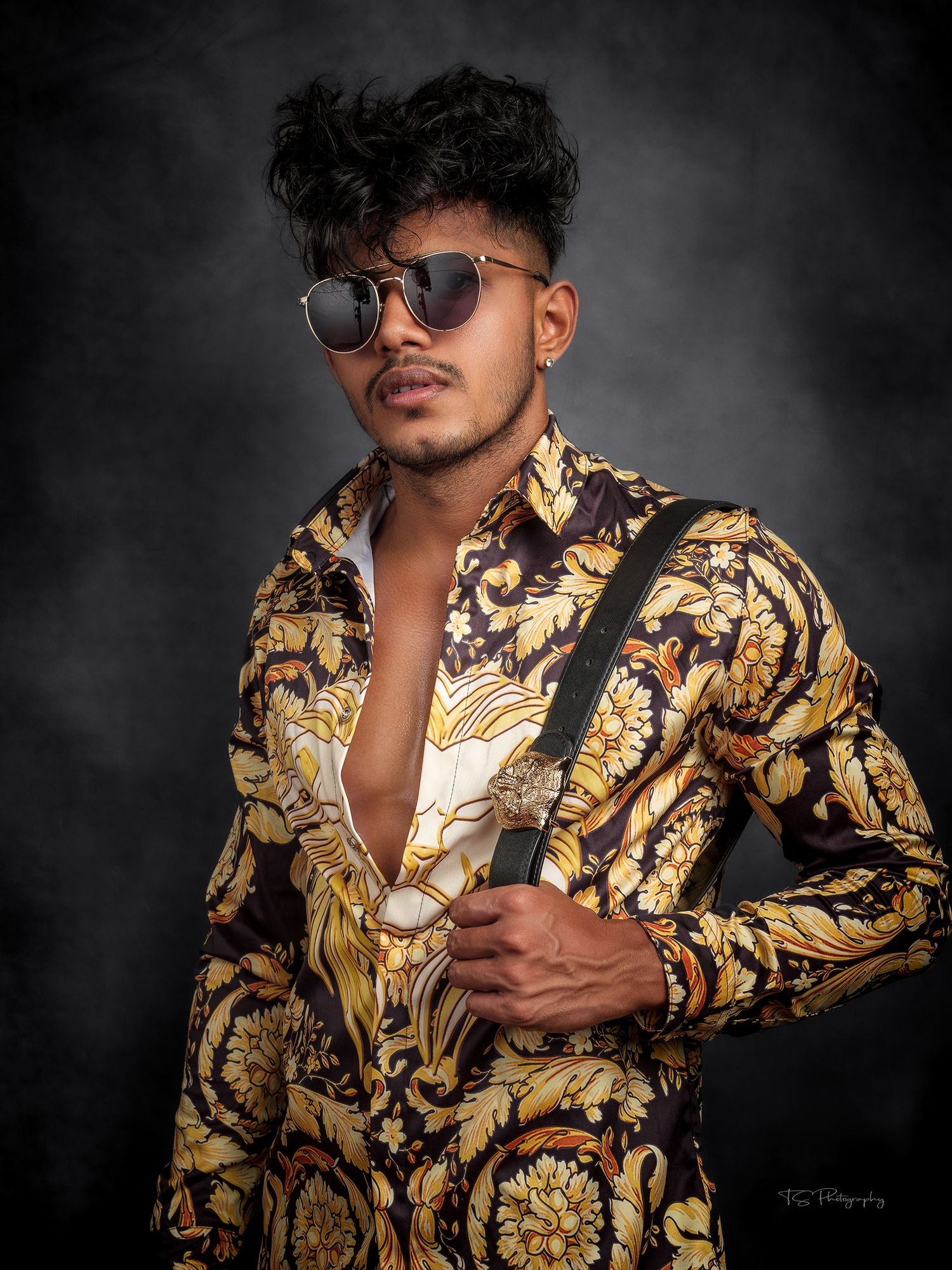 Fashion portrait featuring ornate gold and black patterned shirt with sunglasses against dark background.