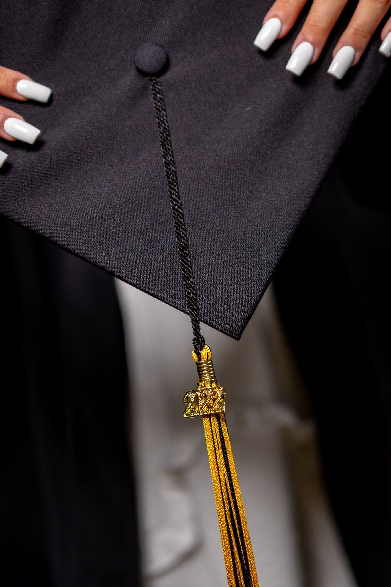 Close-up of graduation cap with white manicured nails and gold tassel against dark background.