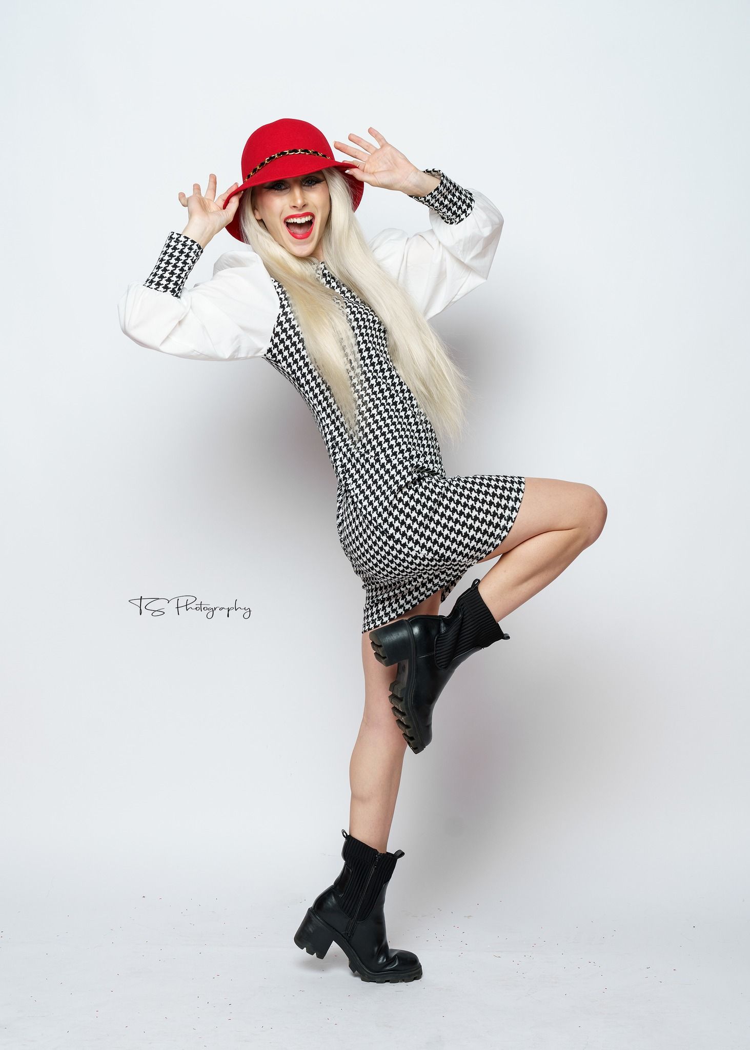 Photograhy, photograpyPlayful fashion model in houndstooth dress and red cap poses energetically with  boots against white background.