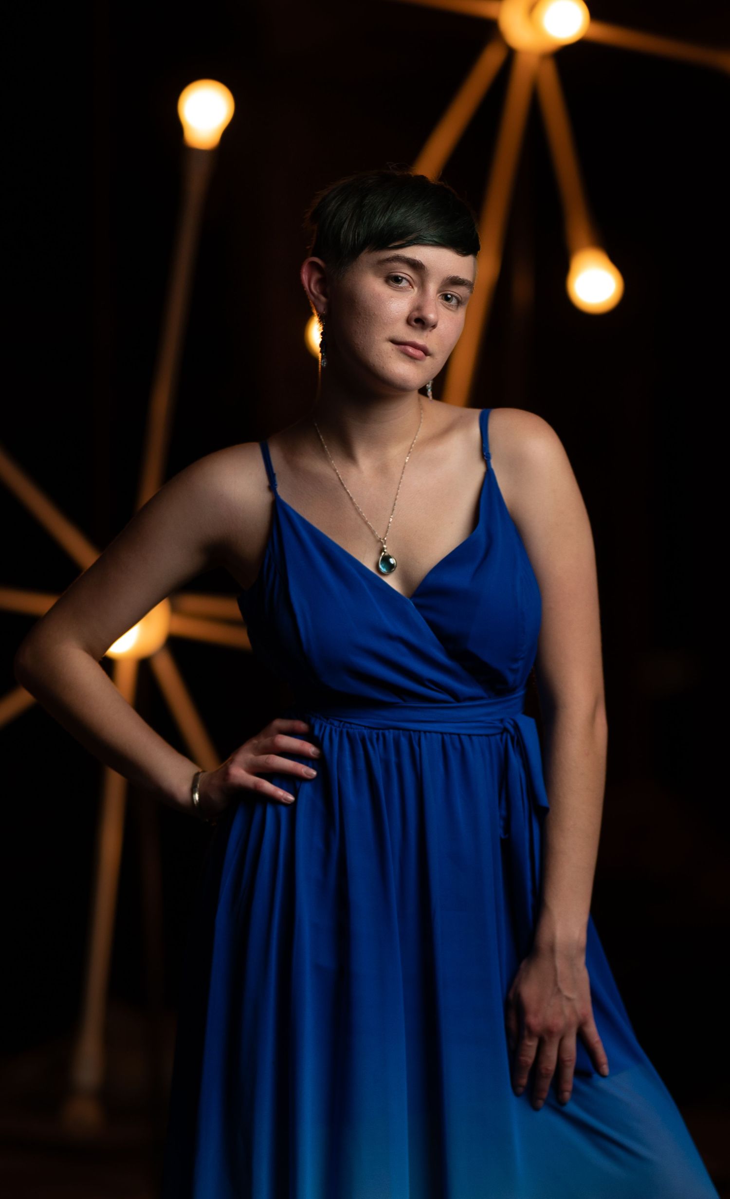 A woman in a royal blue wrap dress poses against a dark background with glowing lights overhead.