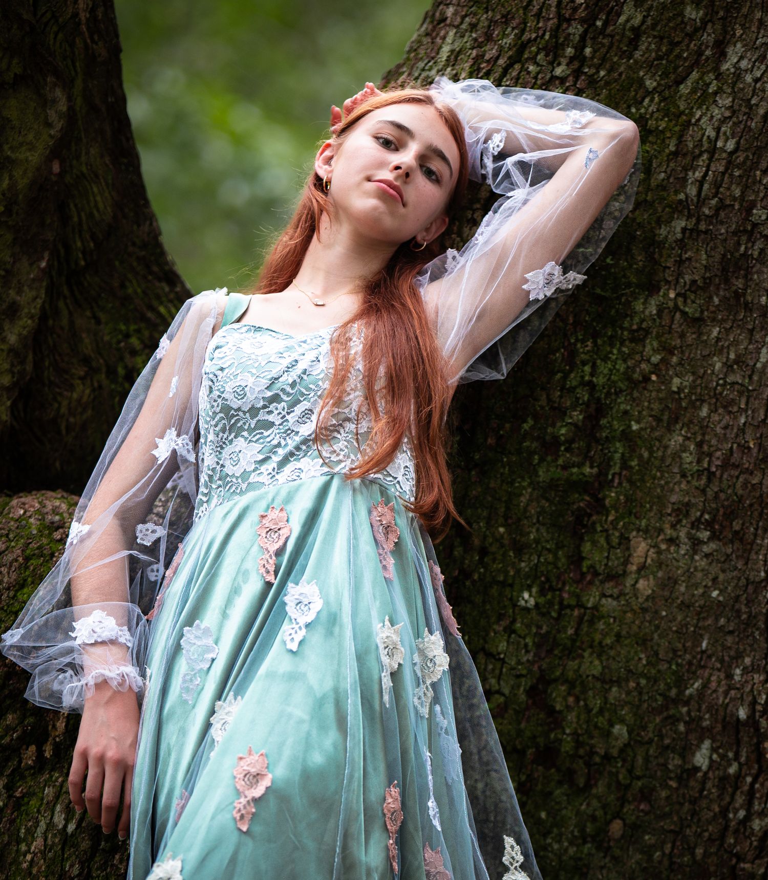 Fantasy-inspired photo of a dreamy ethereal dress in mint blue with sheer sleeves and floral embellishments against a forest tree.
