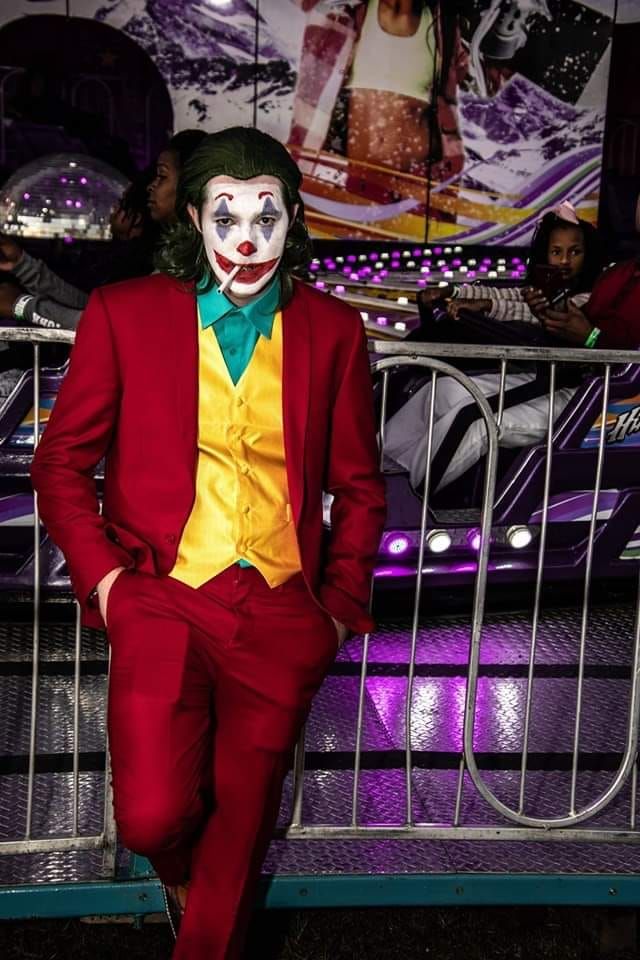 A costumed figure in a red suit and yellow vest poses at a carnival with colorful purple lights in the background.